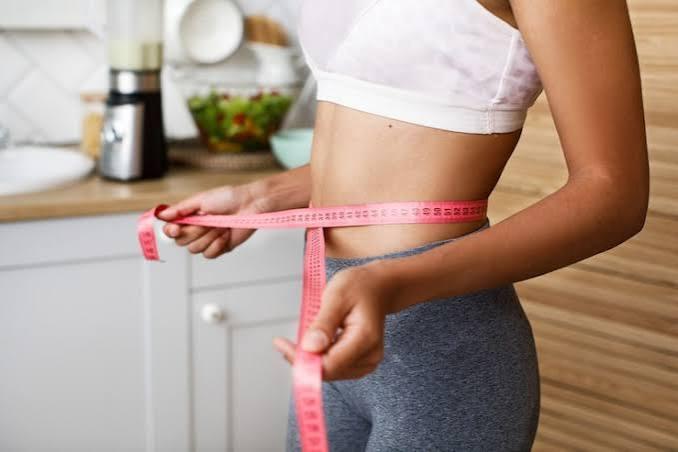 Adult measuring waist in a kitchen while tracking healthy habits